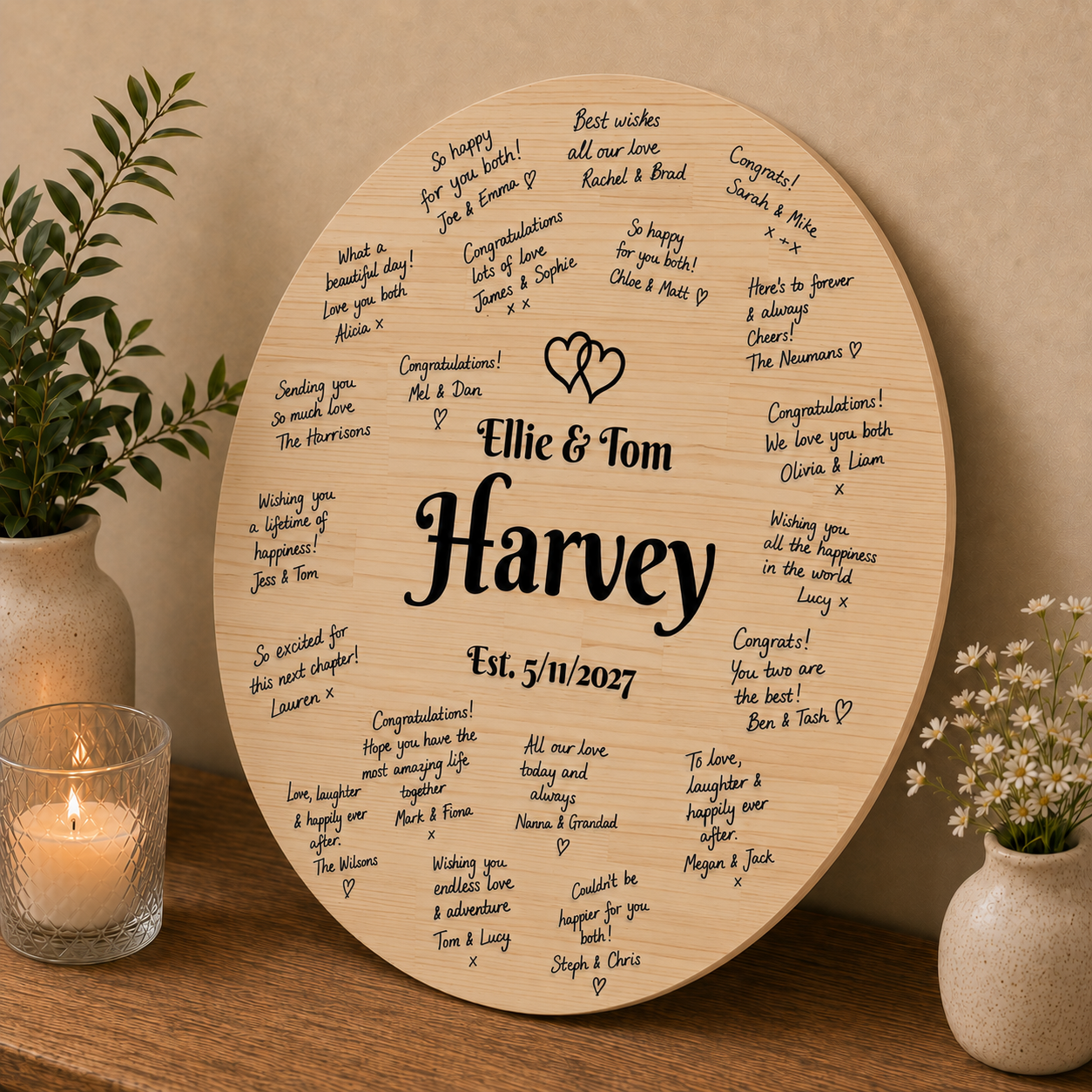 A round wooden personalized gift plaque engraved with 'Elie & Tom Harvey Est. 5/11/2027' and surrounded by handwritten congratulatory messages, styled on a wooden table with a lit candle and small potted plants.